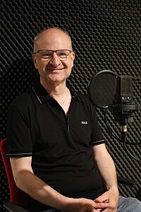 Porträt: Prof. Heilmann im Studio vor einer Wand aus grauem Akustikschaumstoff, vor ihm ein Mikrofon