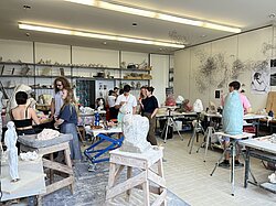 Im Bildhauerei-Atelier: verschiedene Leute, die sich dort aufhalten, zwischen angefangenen und fertiggestellten Skulpturen und Plastiken