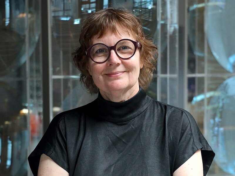Porträtfoto: lächelnde Frau mit rötlichem Kurzhaarschnitt, Brille mit breitem Rahmen und schwarzem Poloshirt vor neutralem Hintergrund