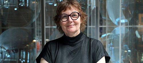 Porträtfoto: lächelnde Frau mit rötlichem Kurzhaarschnitt, Brille mit breitem Rahmen und schwarzem Poloshirt vor neutralem Hintergrund